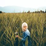 Blonde woman in a denim jacket exploring a verdant field under a bright sky.